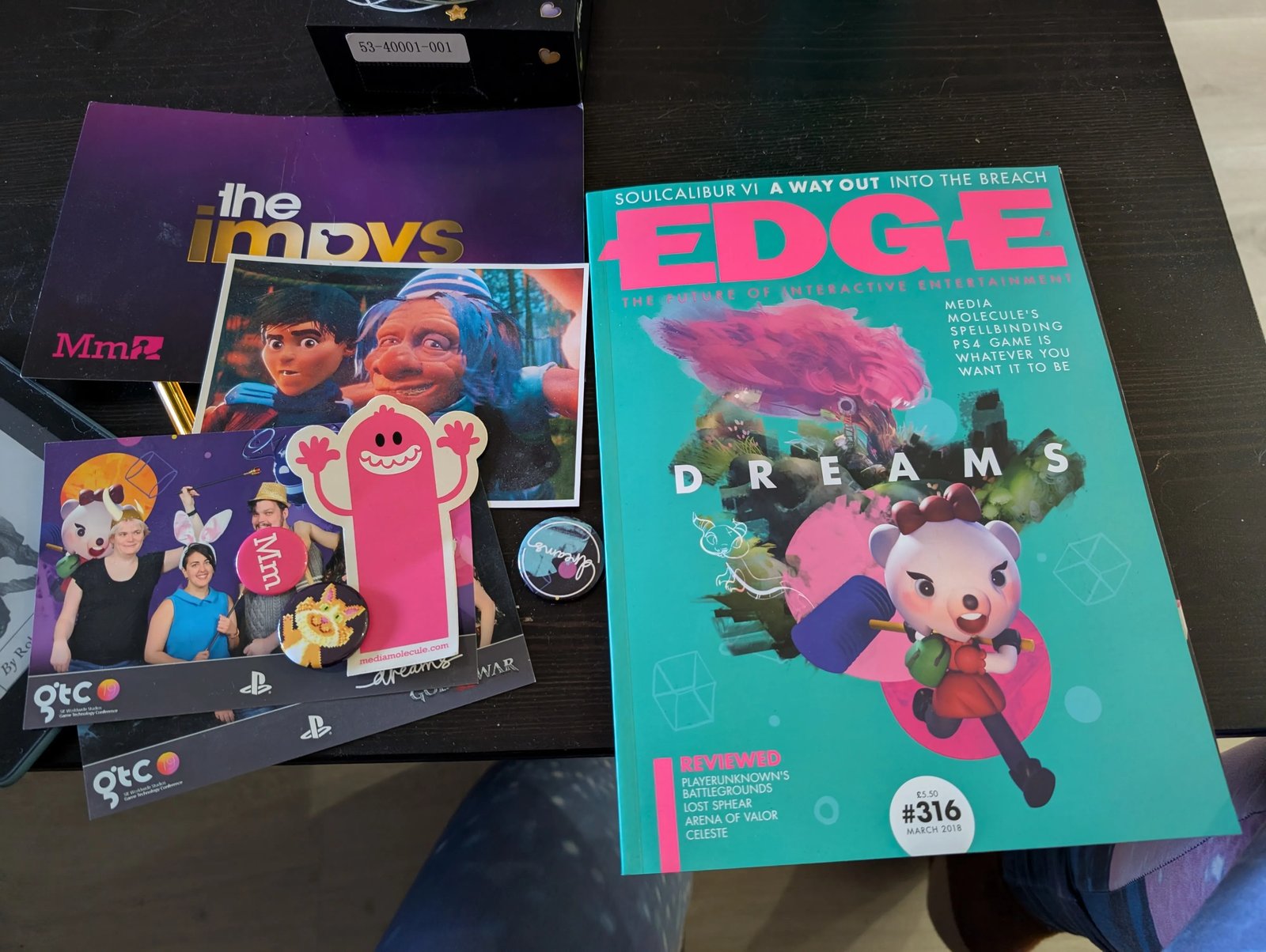 A desk covered in Dreams memorabilia: a purple Impys card with the Mm logo, Edge magazine issue 316 from March 2018 with Dreams on the cover, GTC 19 photobooth prints, a pink Media Molecule sticker, an Mm badge and a Dreams badge