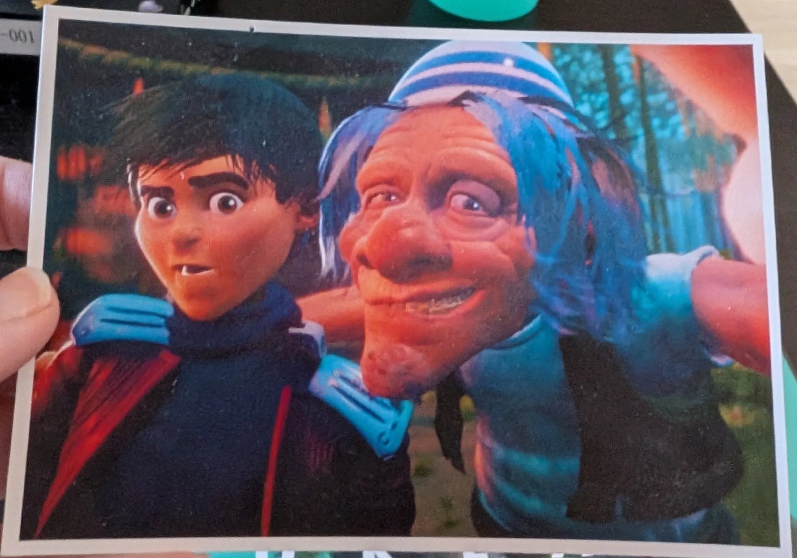 A printed still from Noguchi's Bell, made in Dreams: a wide eyed young man in a dark scarf, and a grinning old man with blue hair and a striped hat with his arm around him