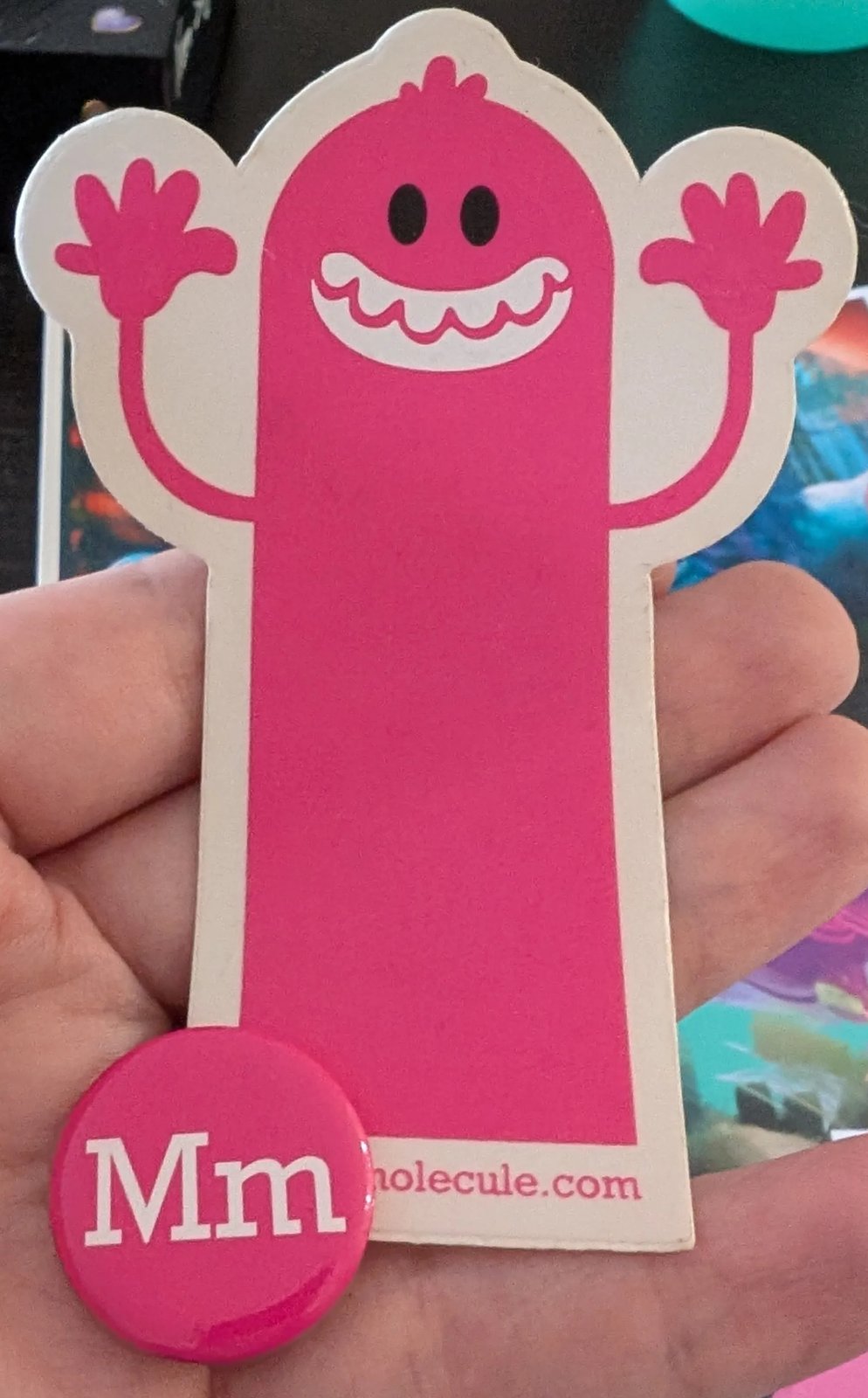 A pink Media Molecule sticker of a tall grinning character with its arms up, and a pink Mm badge