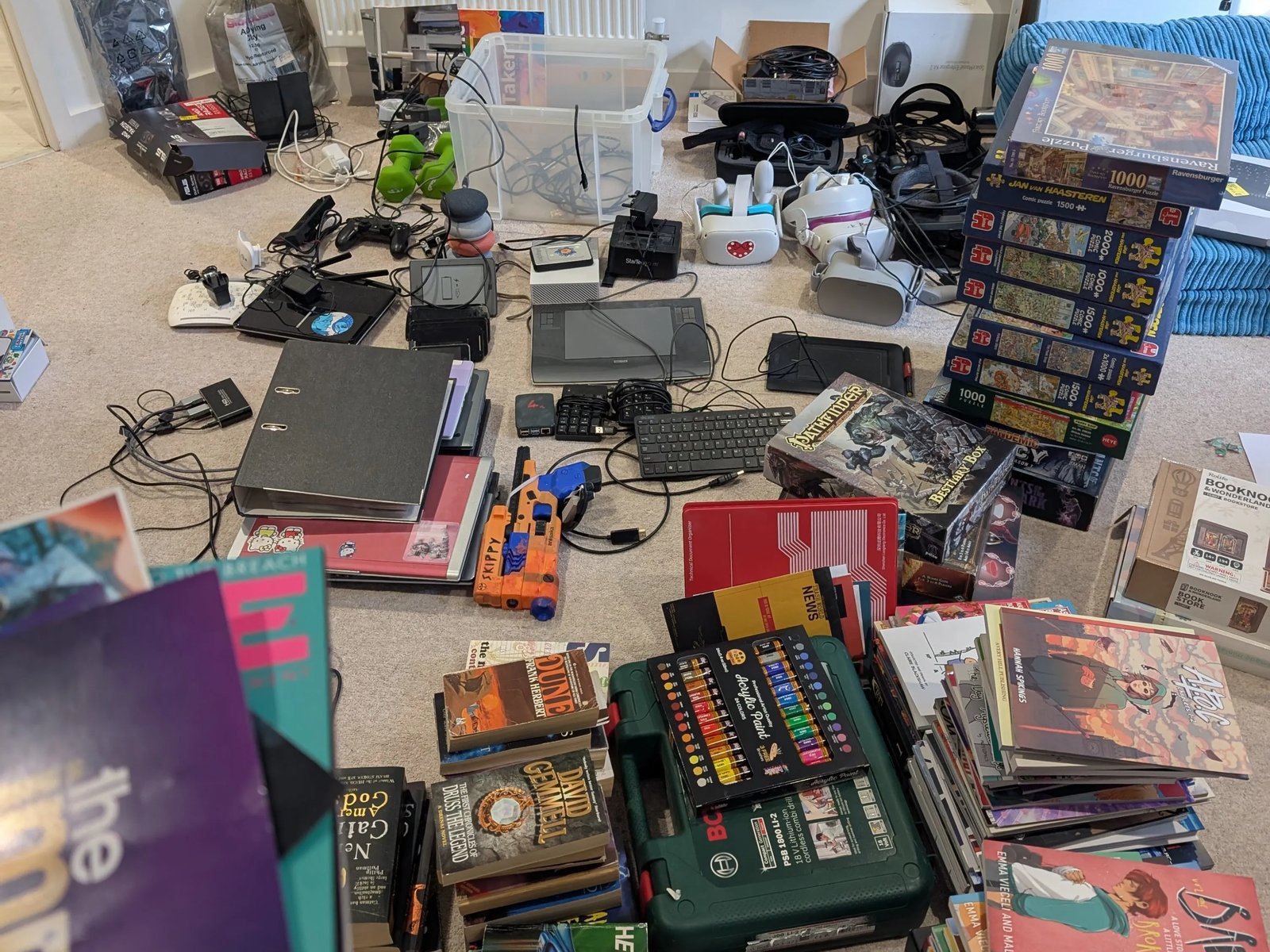The study floor mid sort: piles of books and comics, stacked jigsaw puzzles, VR headsets, drawing tablets, keyboards and cables, with the Impys card in the corner